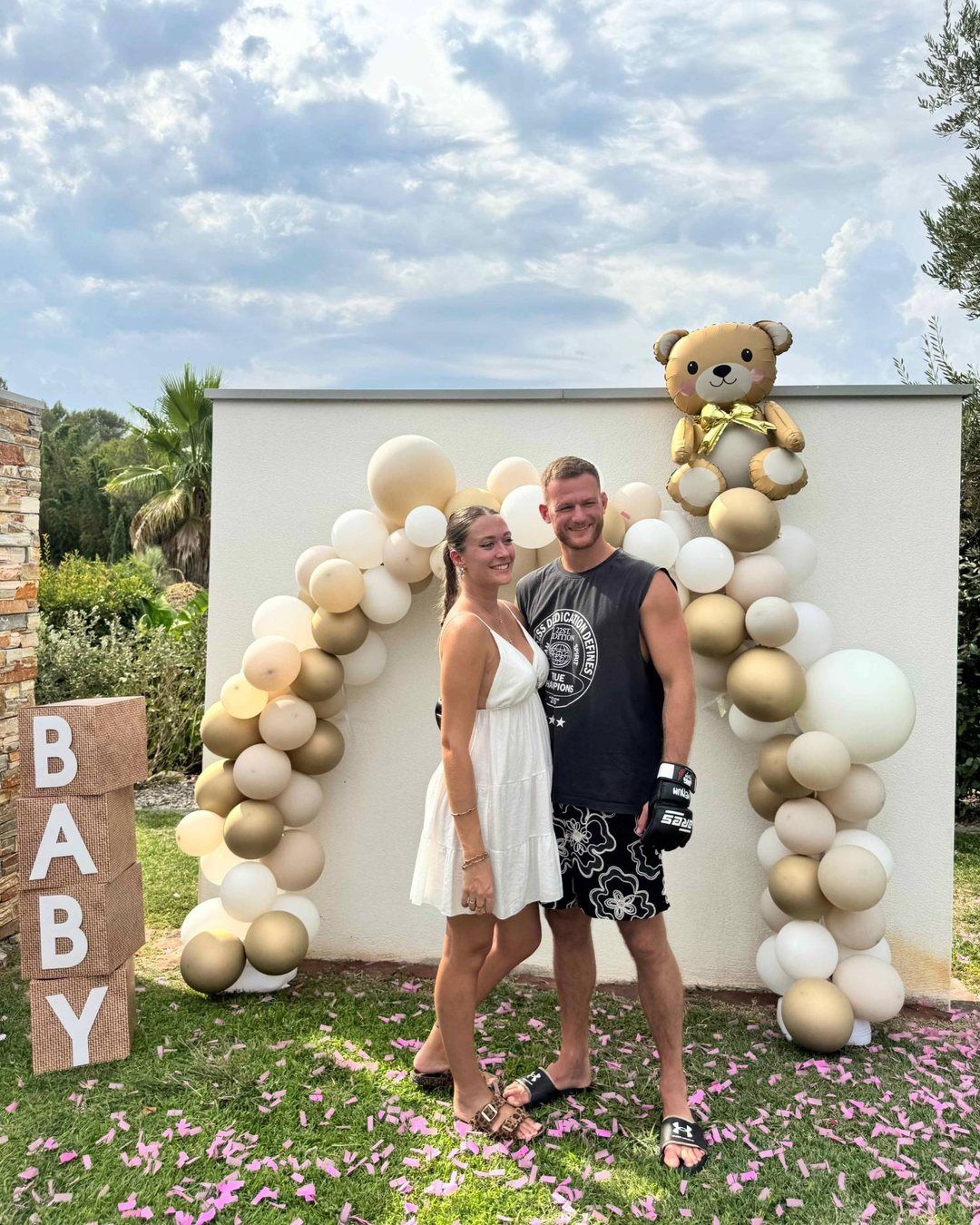 Les futurs parents devant une arche de ballons dorés et crème, un ourson en ballon au-dessus d'eux et des confettis roses au sol